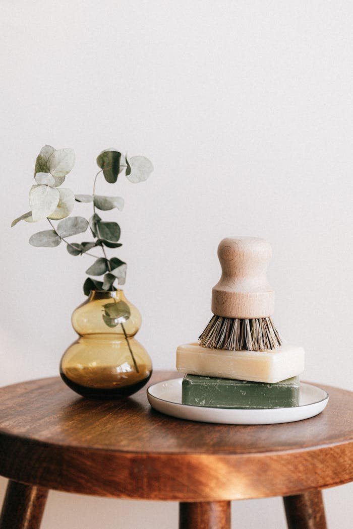Composition of shaving brush placed on organic soaps on ceramic white holder near yellow transparent vase with plant on wooden round table against white wall